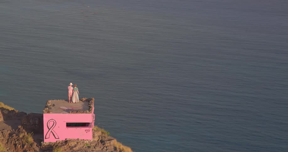 An aerial shot pulls back from two hikers on a pink breast cancer awareness bunker to reveal the stunning coastline and ocean below