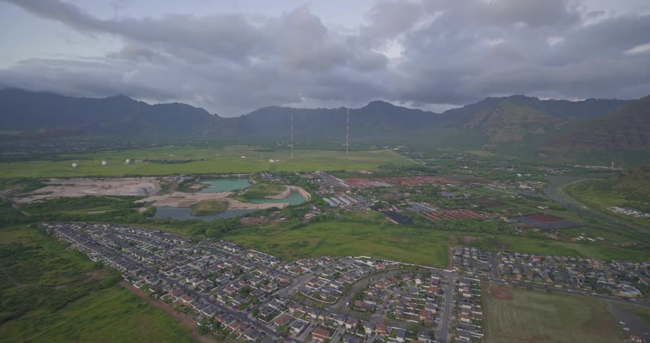 An expansive aerial shot shows a suburban town in a verdant valley flanked by dramatic mountains under an overcast sky