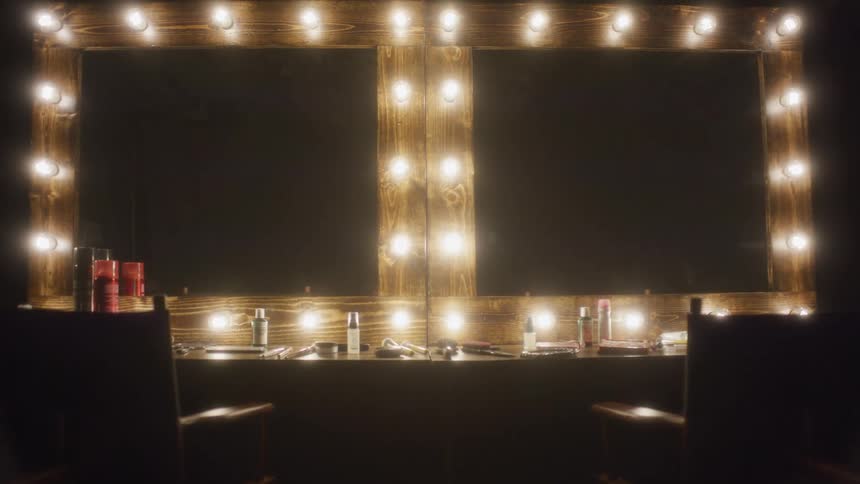 The bright vanity lights surrounding two mirrors in a dark dressing room slowly dim and fade to black