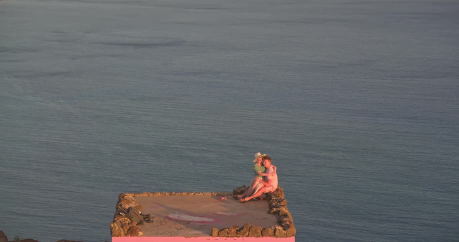 A slow zoom out reveals a romantic couple cuddling on a pink cliffside lookout, admiring the breathtaking ocean and coastal city view at sunset