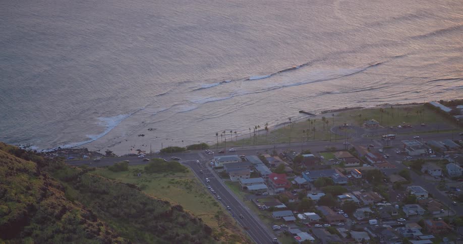 A serene high-angle view of a coastal highway and town nestled between a green hillside and the calm ocean at dusk
