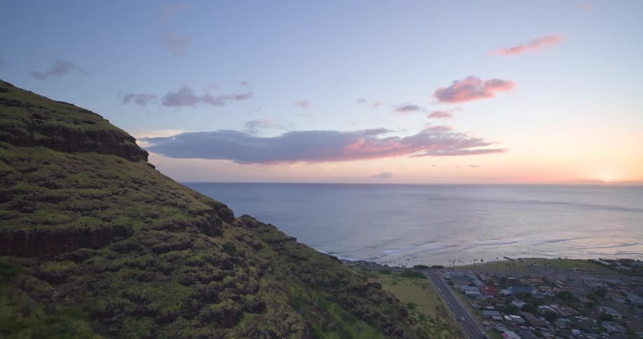 An aerial view ascends a lush green mountain overlooking a coastal highway and town as the sun sets over the pacific ocean