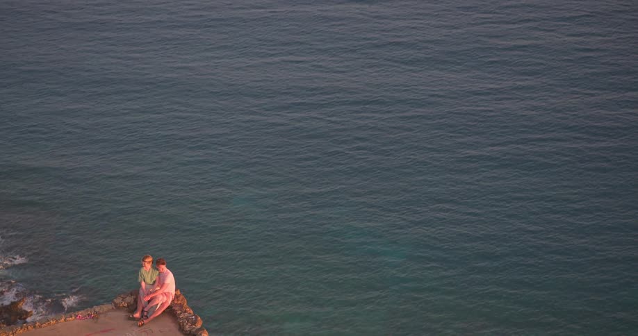 An epic aerial shot pulls back from a young couple sitting on a scenic cliff to reveal the vast blue ocean below