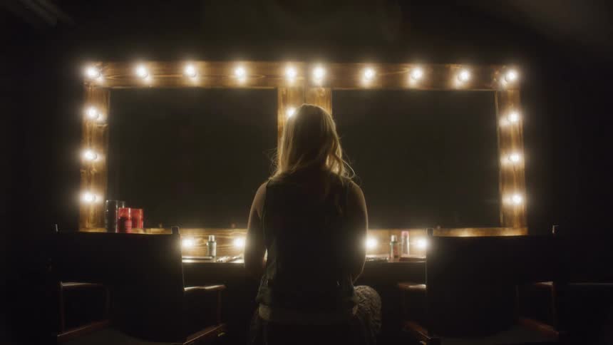 A lone blonde woman sits at a brightly lit vanity mirror in a dark, atmospheric dressing room as the camera slowly pulls back