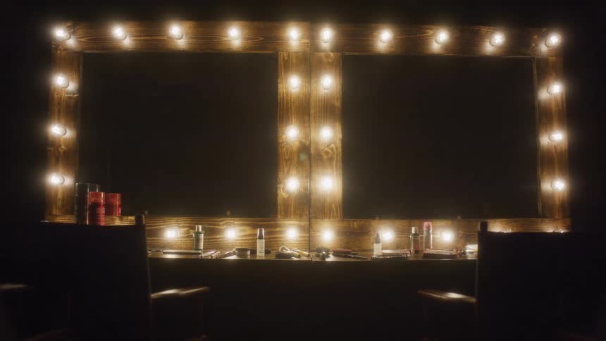 Vanity lights slowly illuminate an empty double makeup station in a dark backstage dressing room