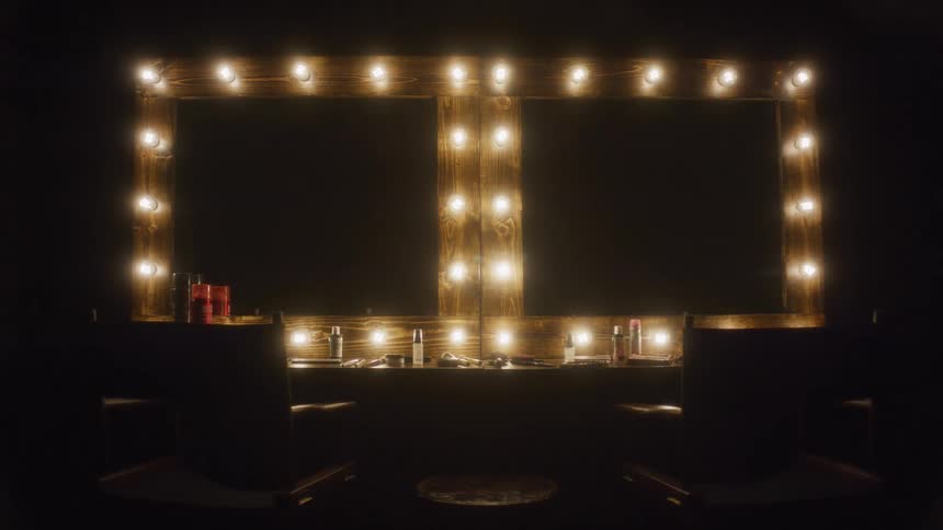 Two empty chairs sit in a dark room facing a large makeup mirror framed with glowing bulbs