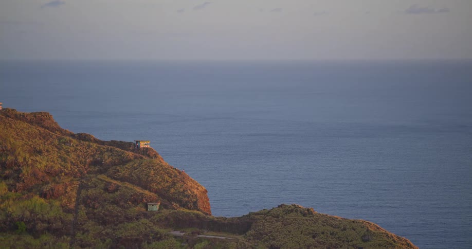A slow zoom out reveals old military bunkers and small houses on a lush, golden-lit coastal hill overlooking the calm ocean