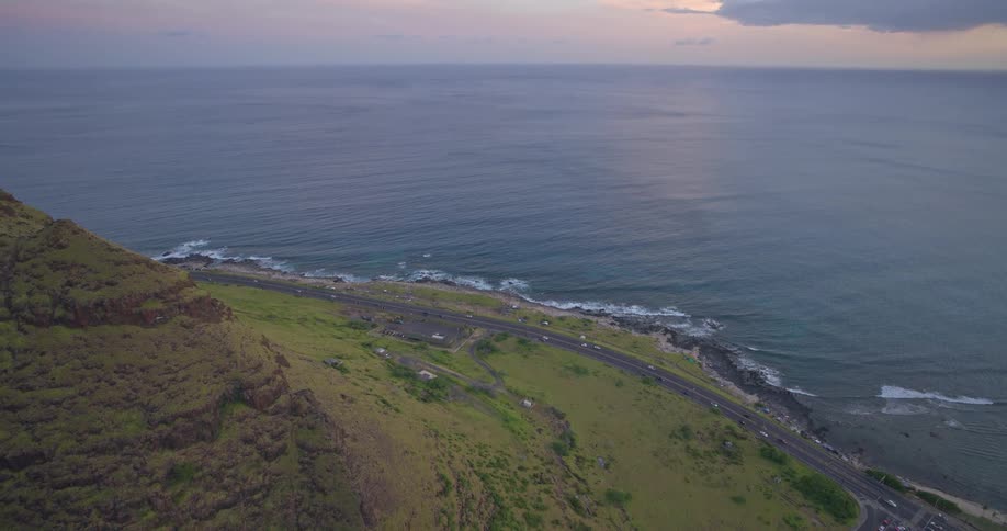 A stunning aerial view pulls back to reveal a dramatic green mountain cliffside overlooking a scenic coastal highway and the tranquil ocean at sunset