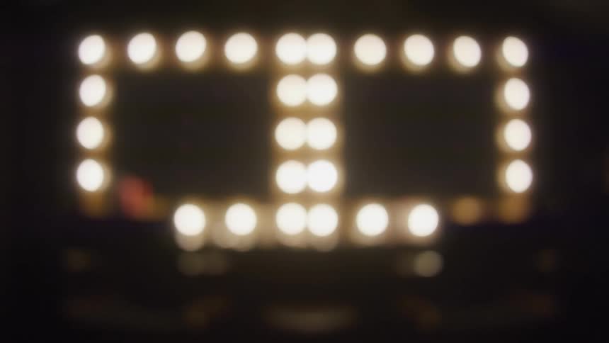 A slow zoom out from the blurred bokeh lights of a backstage makeup vanity mirror in a dark room