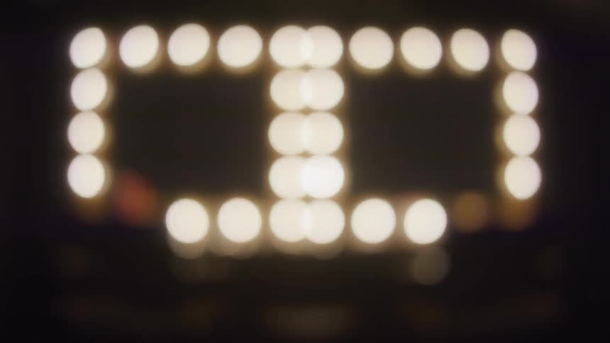 An empty backstage dressing room with two brightly lit vanity mirrors slowly goes out of focus