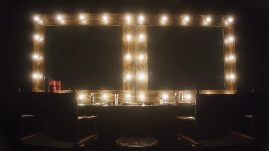 A classic backstage makeup vanity with two mirrors is revealed as the surrounding light bulbs slowly turn on in a dark room