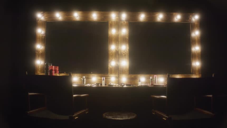 An empty backstage makeup station is slowly revealed as the vanity mirror lights turn on and then fade back to black