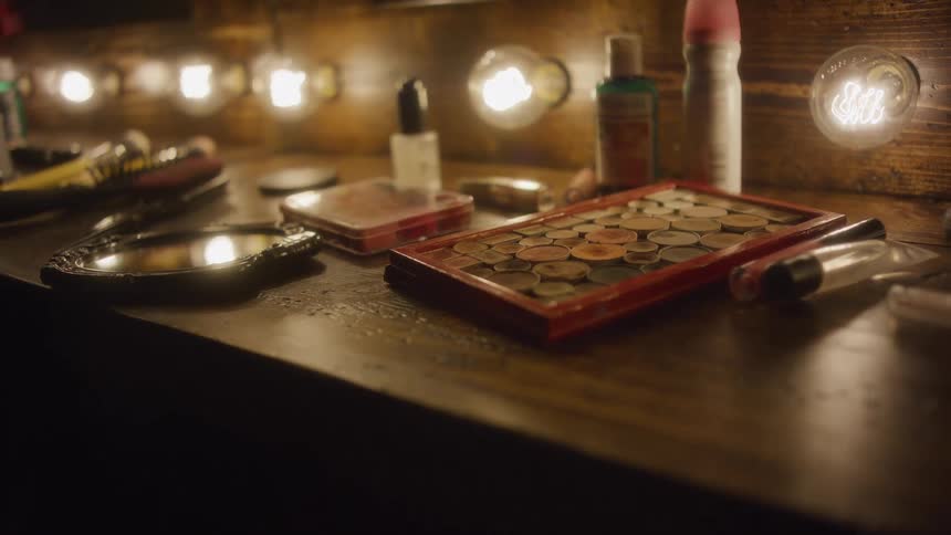 A slow, intimate pan across a rustic wooden makeup vanity table cluttered with cosmetics under the warm glow of vintage light bulbs