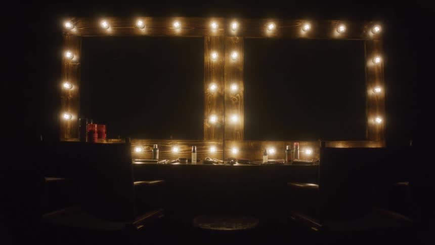 A wooden double vanity mirror with light bulbs slowly illuminates an empty backstage dressing room