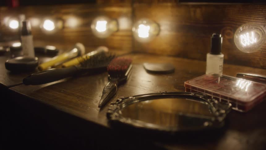 A slow pull-back shot reveals various cosmetic products on a rustic wooden makeup vanity with warm vintage light bulbs