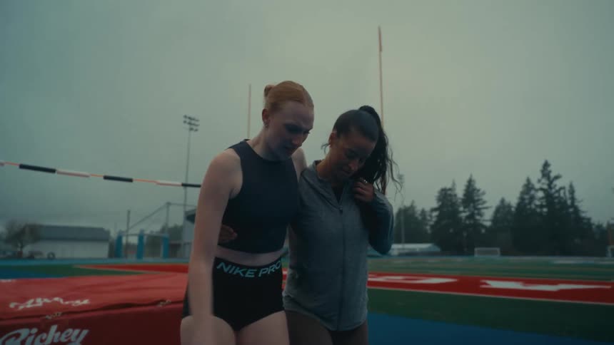 An injured female track and field athlete is supported by her teammate as they walk off the field together