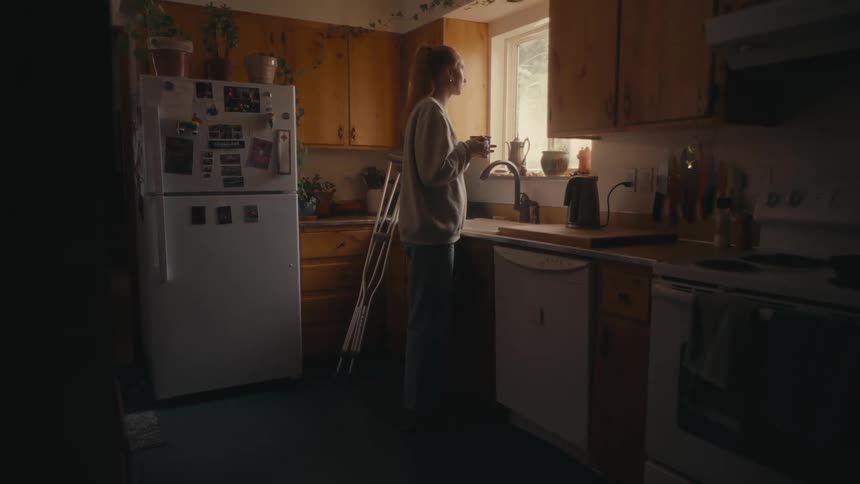 A pensive young woman holding a mug gazes out the kitchen window in the morning light, with crutches resting nearby
