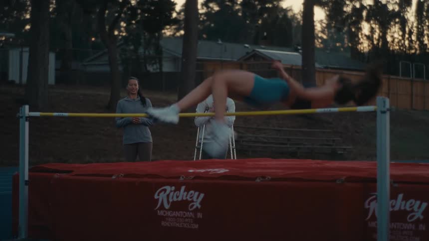 A female high jumper gracefully clears the bar in slow motion as her coach and an injured teammate watch and clap