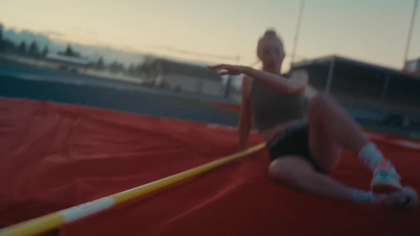 A young female athlete successfully clears the bar in a slow-motion high jump, landing safely on the red mat at an outdoor track