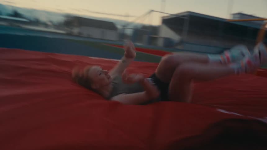 A female athlete makes a successful high jump in slow motion, landing gracefully on the mat at a track and field stadium at dusk