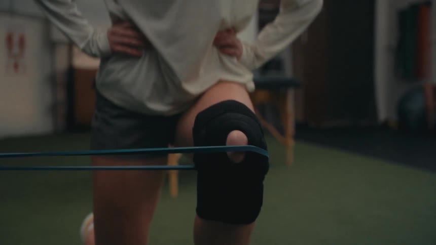 An athlete rehabilitates a knee injury using a resistance band and a supportive brace in a dimly lit gym