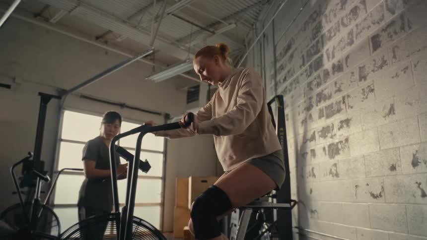 A determined athletic woman performs a cardio workout on an air bike with her trainer watching in a gritty gym