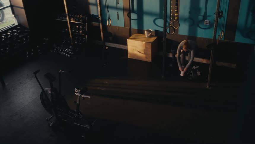 A high-angle view shows a focused woman tying her shoes in a sunbeam before her workout in an empty, dark gym