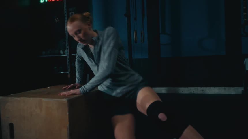 A woman performs a slow-motion box jump in a dark gym and then sits down to rest