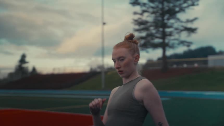 A young female athlete with red hair stretches on a track and field at dusk