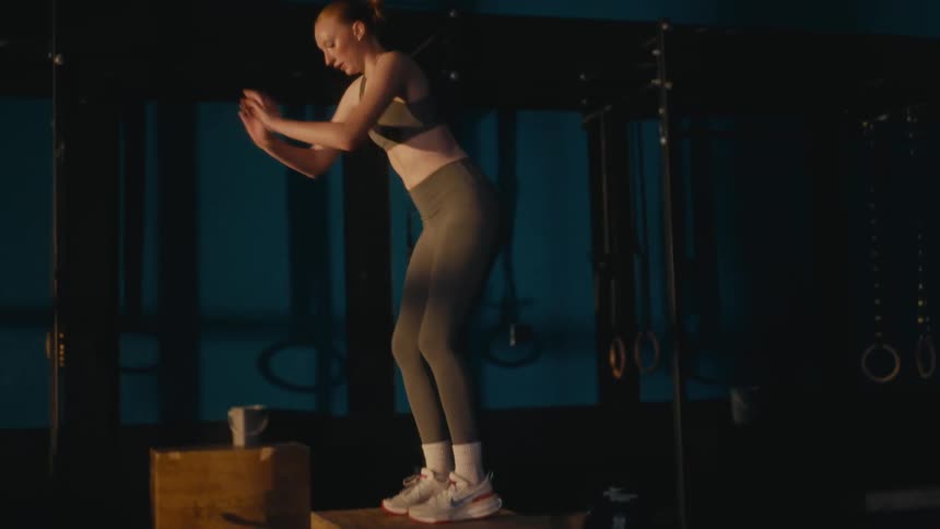 A determined young woman with red hair performs a powerful box jump in slow motion in a dimly lit gym