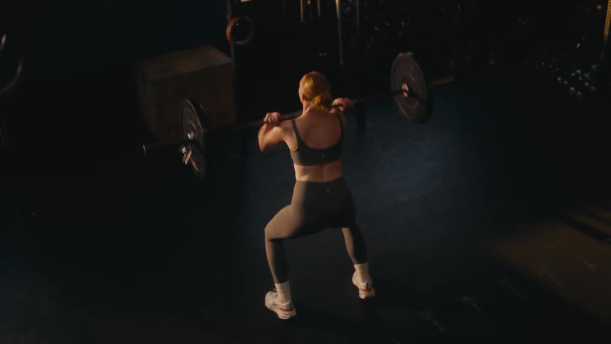 A strong woman from a high angle view does a power clean with a barbell in a dark gym