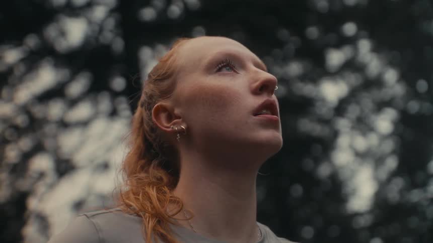 A young woman with red hair takes a deep breath of fresh air and looks up at the sky while standing in a forest
