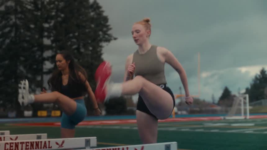 Two female track athletes perform high kick warm up drills over hurdles in slow motion on a cloudy day