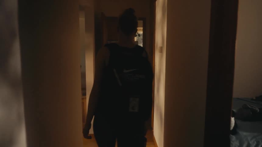 A young woman with a backpack walks down a dimly lit hallway in her home in slow motion