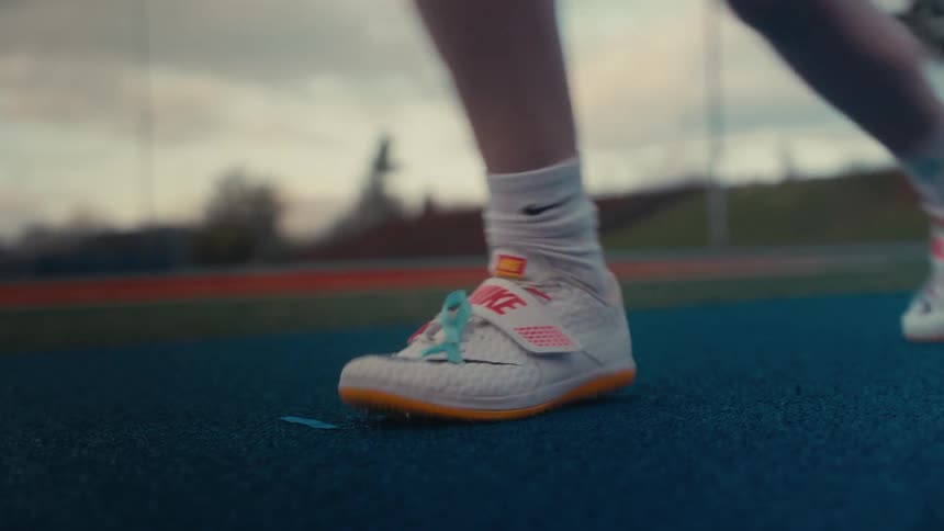 An athlete wearing spiked Nike throwing shoes warms up on a blue track in slow motion