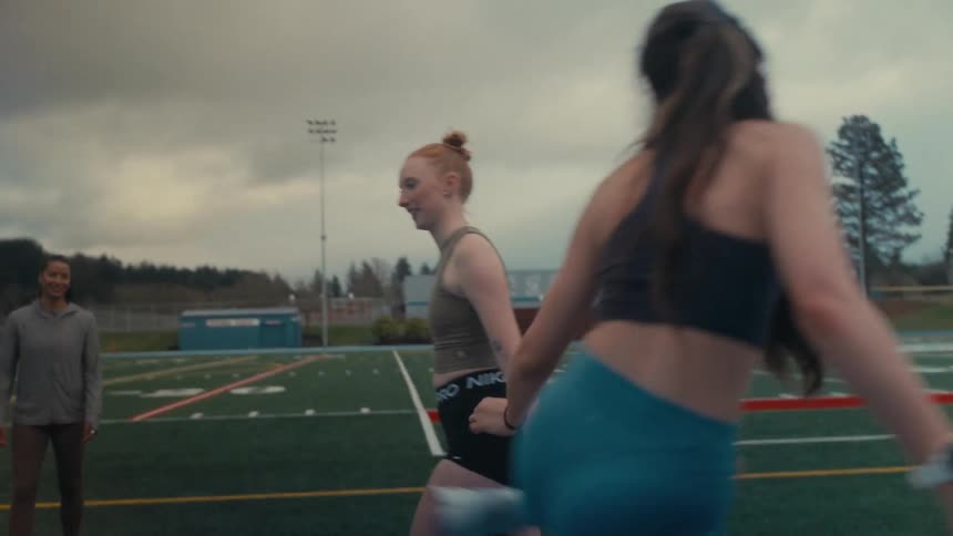 A young woman with red hair stretches and warms up in slow motion with teammates on an athletic field under a cloudy sky