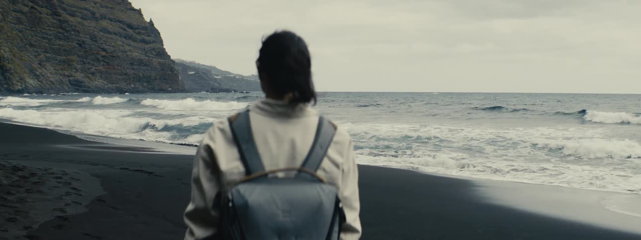 A woman with a backpack stands on a remote black sand beach, watching the ocean waves crash on the shore in slow motion