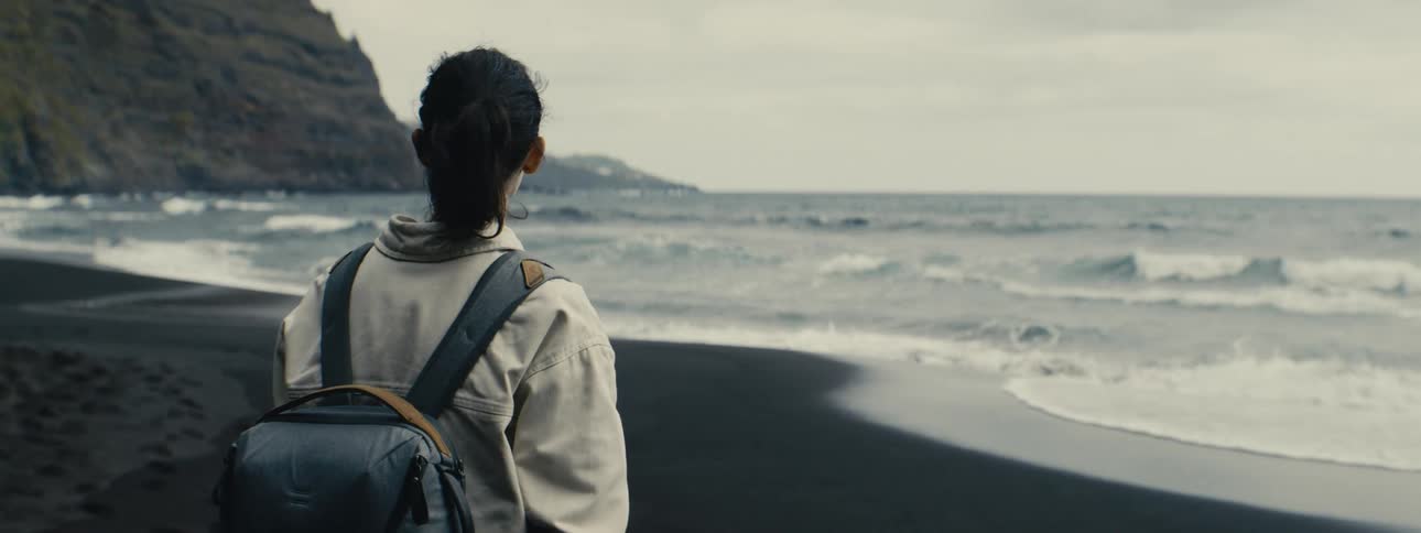 A solo female traveler with a backpack stands on a black sand beach watching the ocean waves roll in