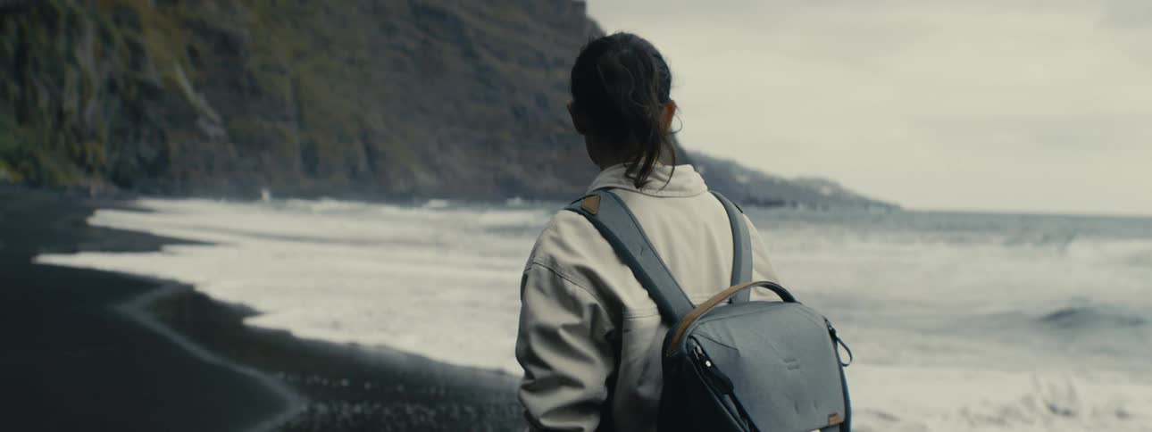 A thoughtful traveler with a backpack looks out at the ocean from a secluded, rugged beach on an overcast day