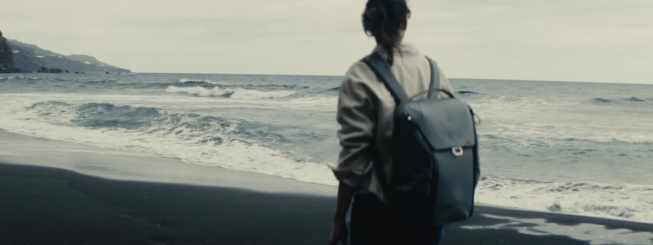 A solitary woman with a backpack stands on a black sand beach watching the waves roll in, filmed in slow motion