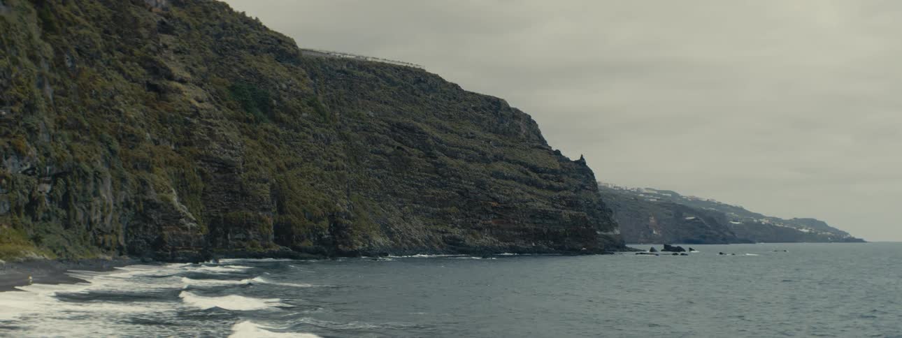 A moody and dramatic wide shot captures a rugged rocky coastline under a grey, overcast sky
