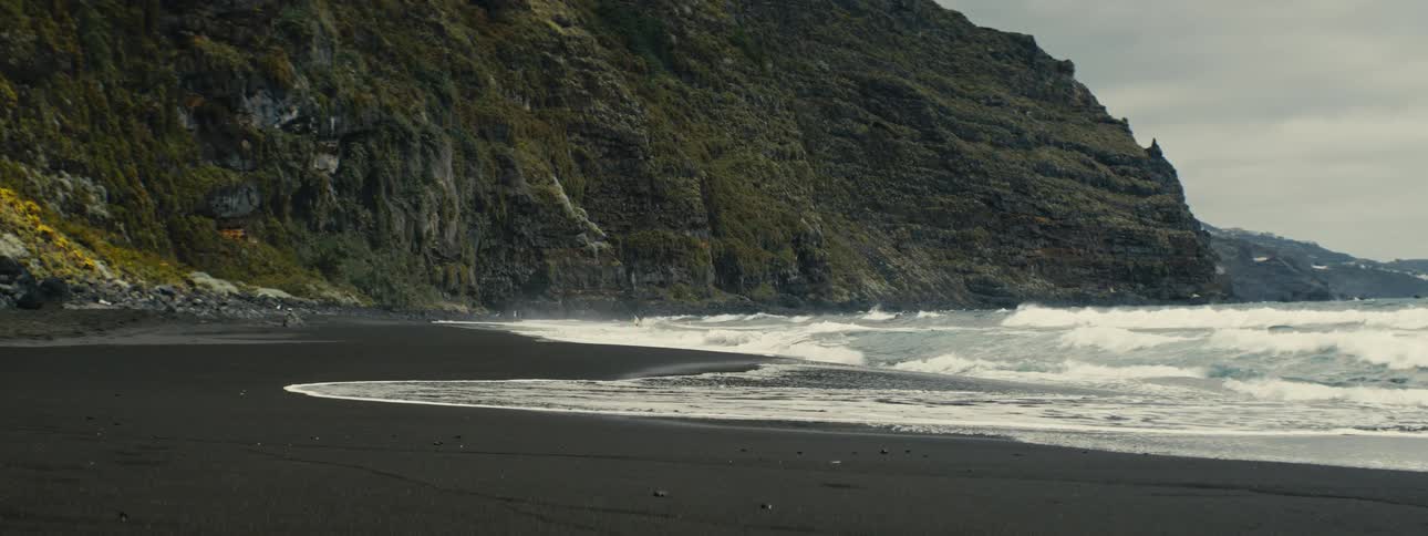 A serene view of gentle ocean waves washing ashore on a black sand beach at the base of a lush green cliff