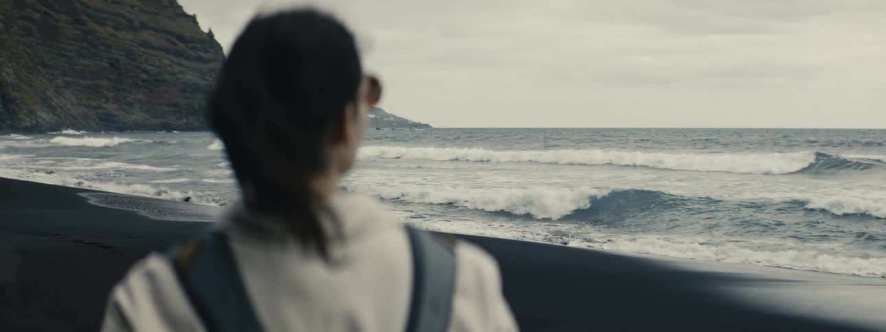 Over-the-shoulder shot of a woman with a windswept ponytail watching the slow-motion waves crash on a moody black sand beach