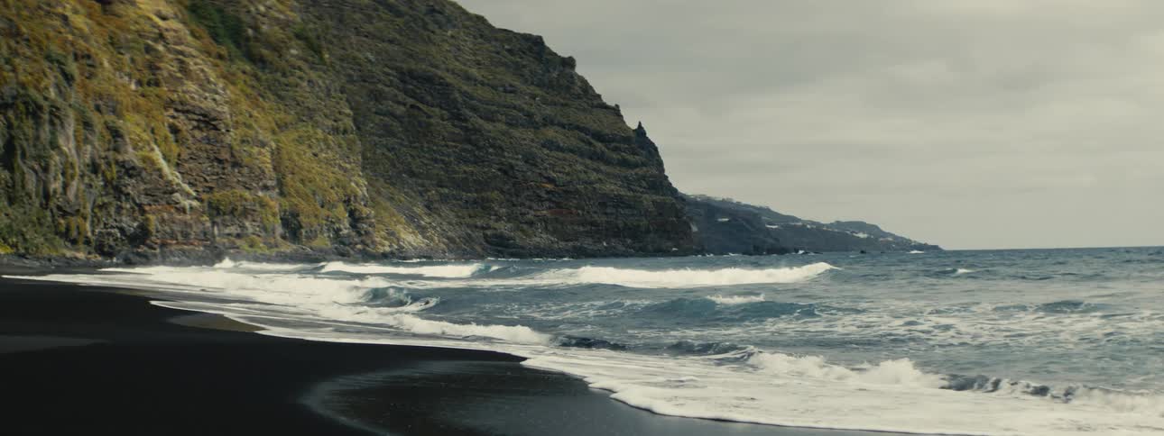 Waves wash ashore in slow motion on a moody black sand beach at the base of a dramatic cliff