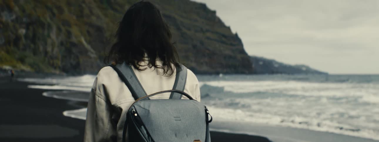 A solo traveler with a backpack walks along a remote black sand beach, observing the ocean waves crashing on the shore