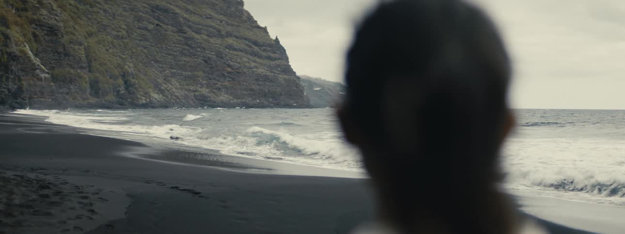 A contemplative slow-motion view from behind a person watching ocean waves roll onto a secluded volcanic black sand beach