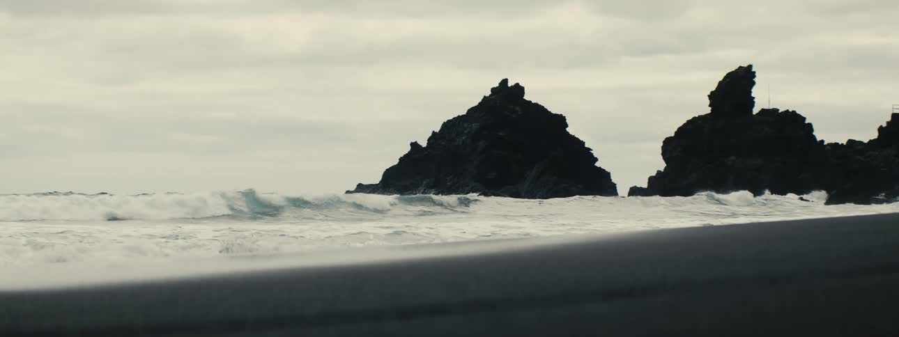 Moody slow-motion waves wash onto a desolate beach in front of a jagged rock formation on a cloudy day