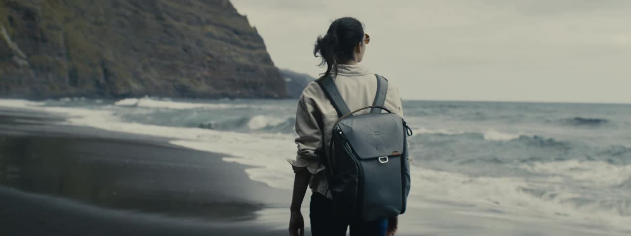 A solo adventurer with a backpack strolls along a moody black sand beach as ocean waves crash on the shore in slow motion