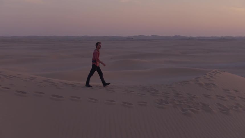 A man with a camera walks in a vast, sandy landscape at dusk