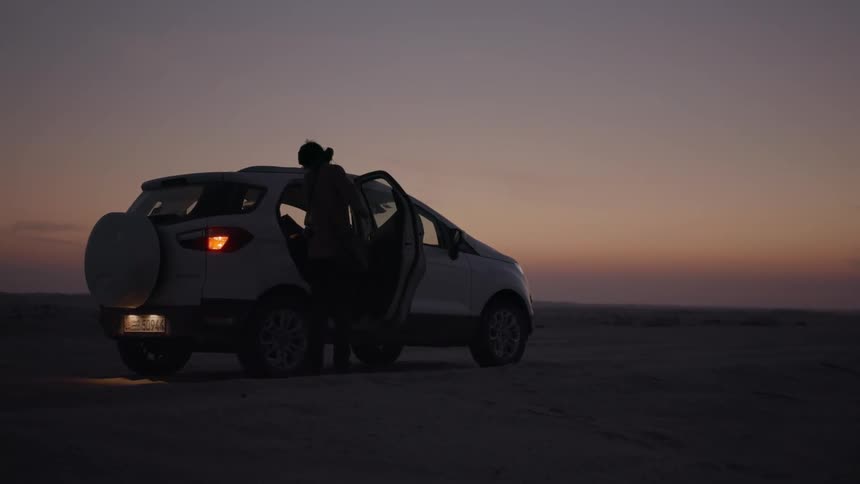 A man approaches a car parked in a desert at sunset